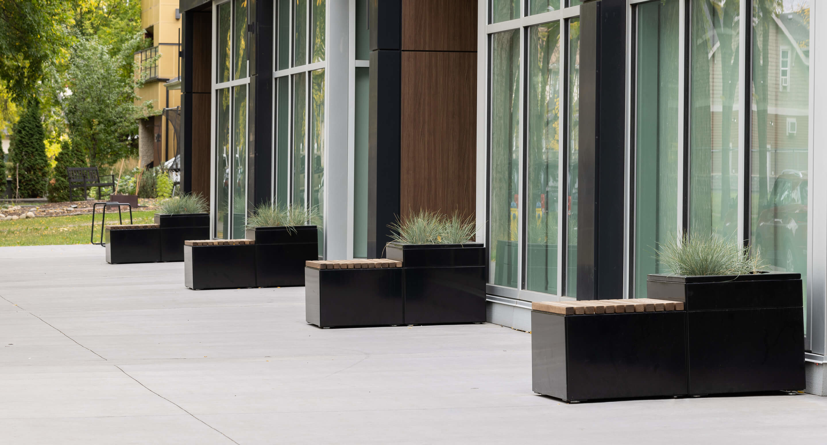 maglin_pixel-planters_2 A row of black planter benches is arranged along the large glass windows of a modern building, with trees and a chair visible in the background.