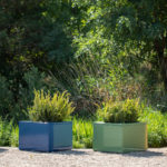 Four square planters in blue, navy, green, and yellow hold green plants, arranged in a row on gravel with leafy trees in the background.