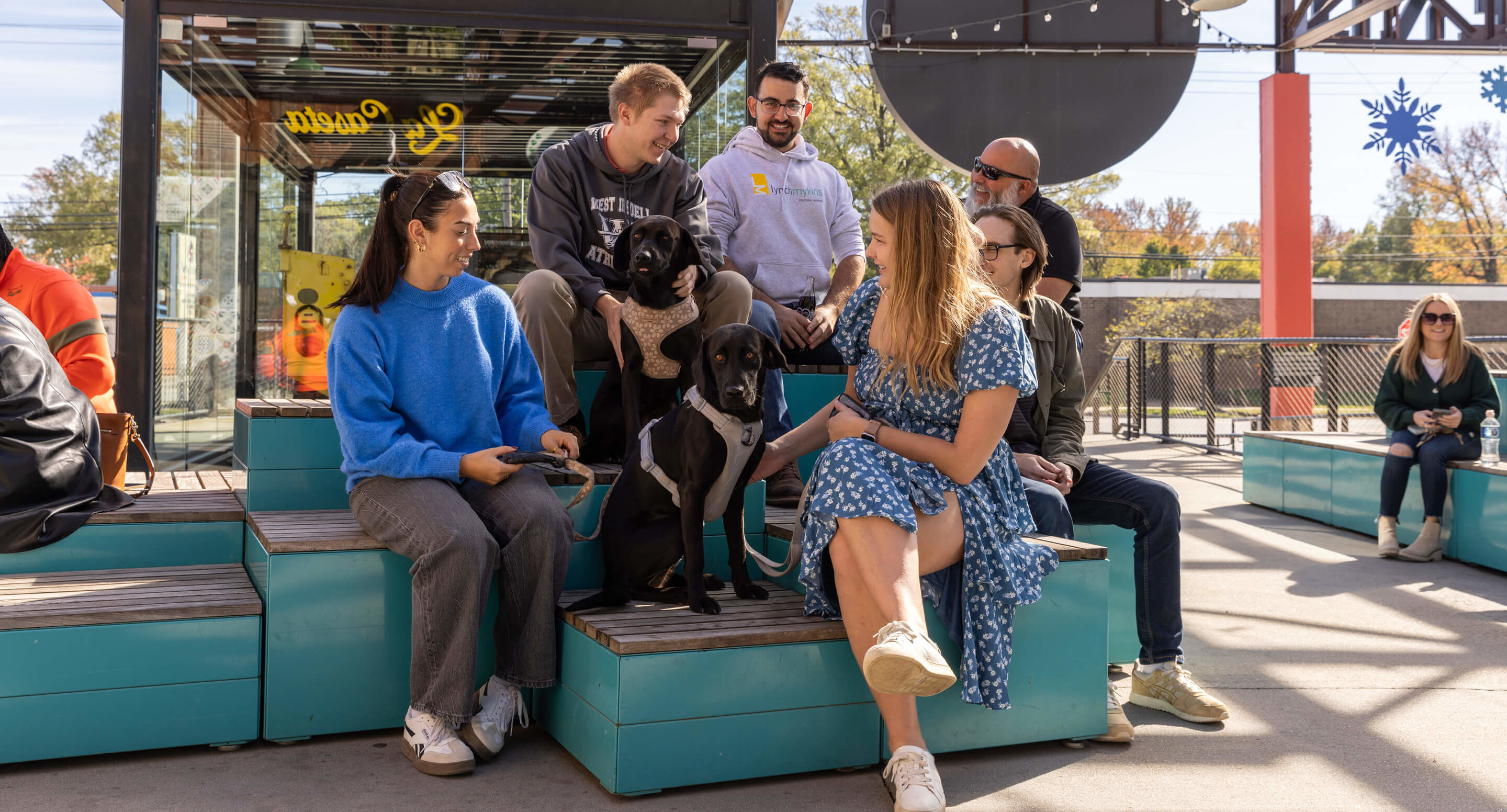 maglin_pixel_1 A group of people sit on tiered benches outdoors with two black dogs, talking and smiling on a sunny day.