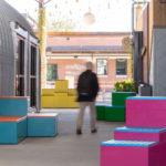 A person walks through an outdoor corridor with colorful square benches in pink, blue, and yellow beside modern buildings.