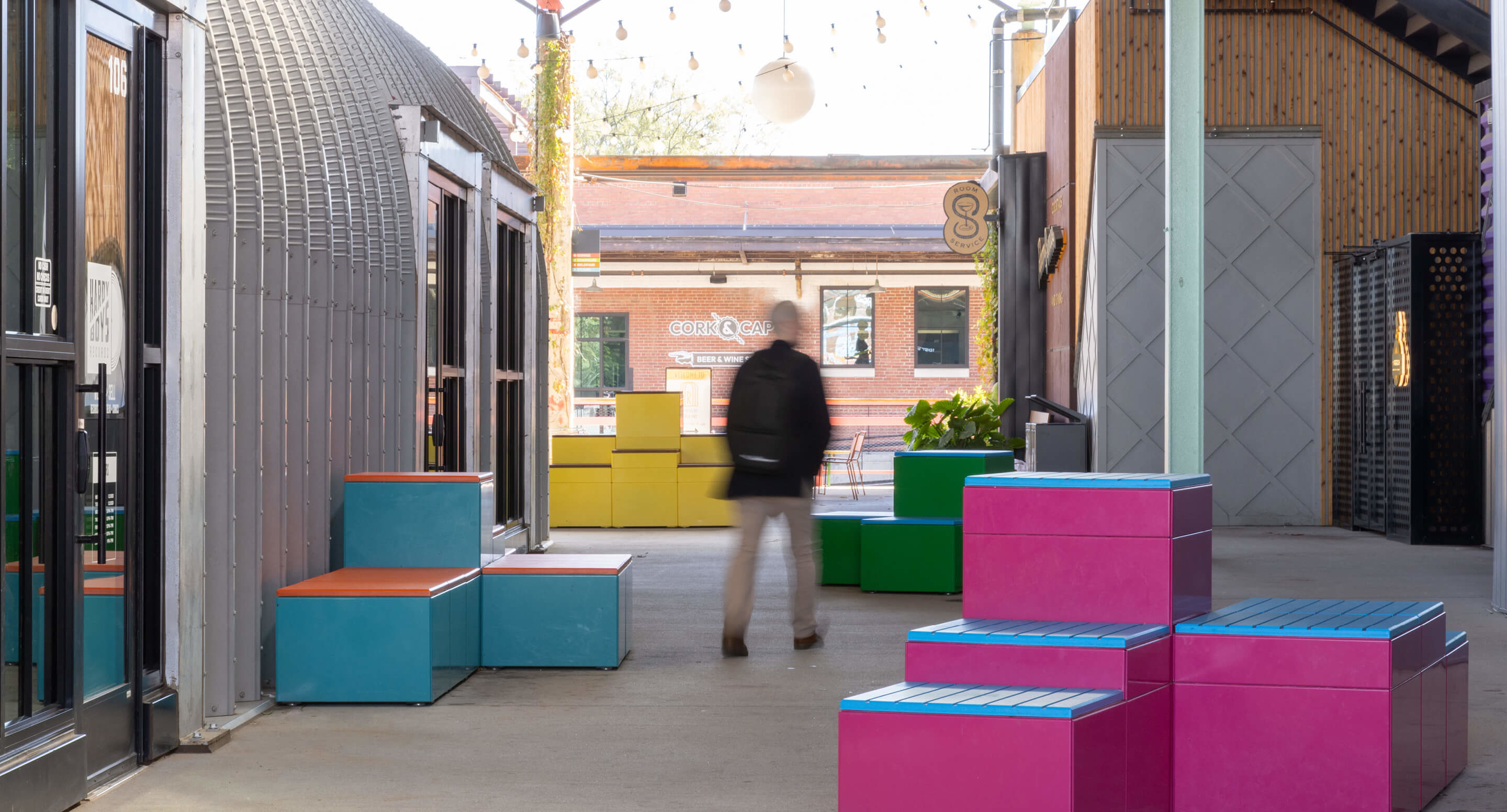 maglin_pixel_2 A person walks through an outdoor corridor with colorful square benches in pink, blue, and yellow beside modern buildings.