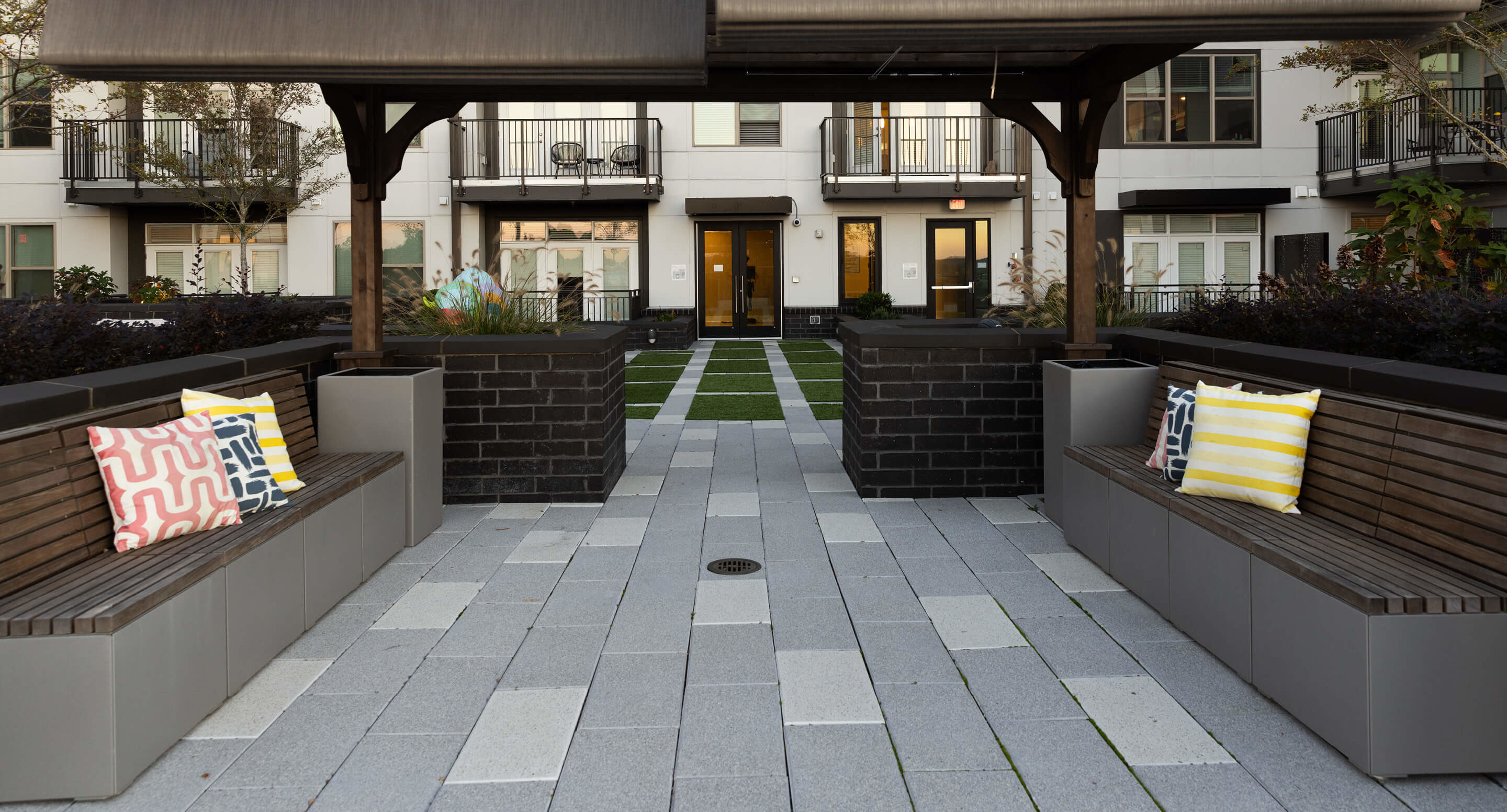 maglin_pixel_3 Outdoor seating area with two wooden benches under a canopy, each with colorful pillows, facing a landscaped courtyard and modern apartment building in the background.