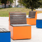 Four modern outdoor benches with wooden slats and bright blue and orange cube bases are arranged on a paved area in a park setting.
