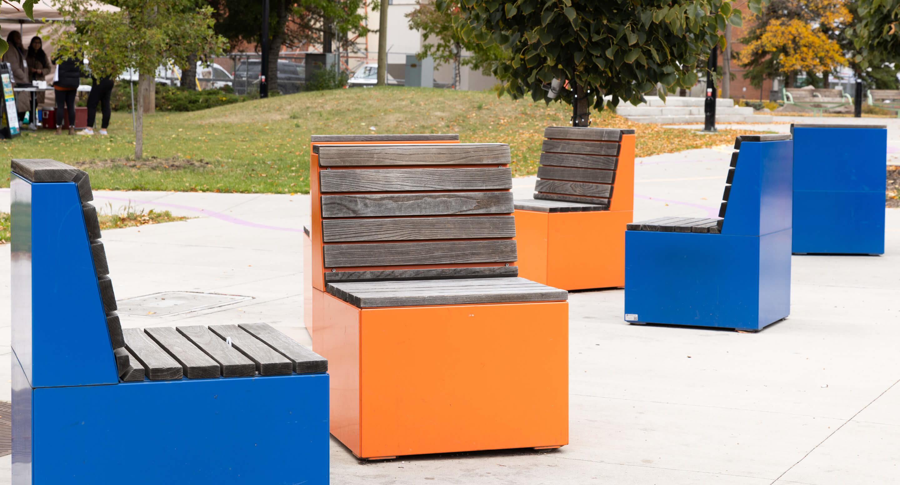 maglin_pixel_4 Four modern outdoor benches with wooden slats and bright blue and orange cube bases are arranged on a paved area in a park setting.