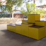 A modular yellow bench structure with wooden seating is positioned outdoors in a shaded area with trees and scattered chairs.