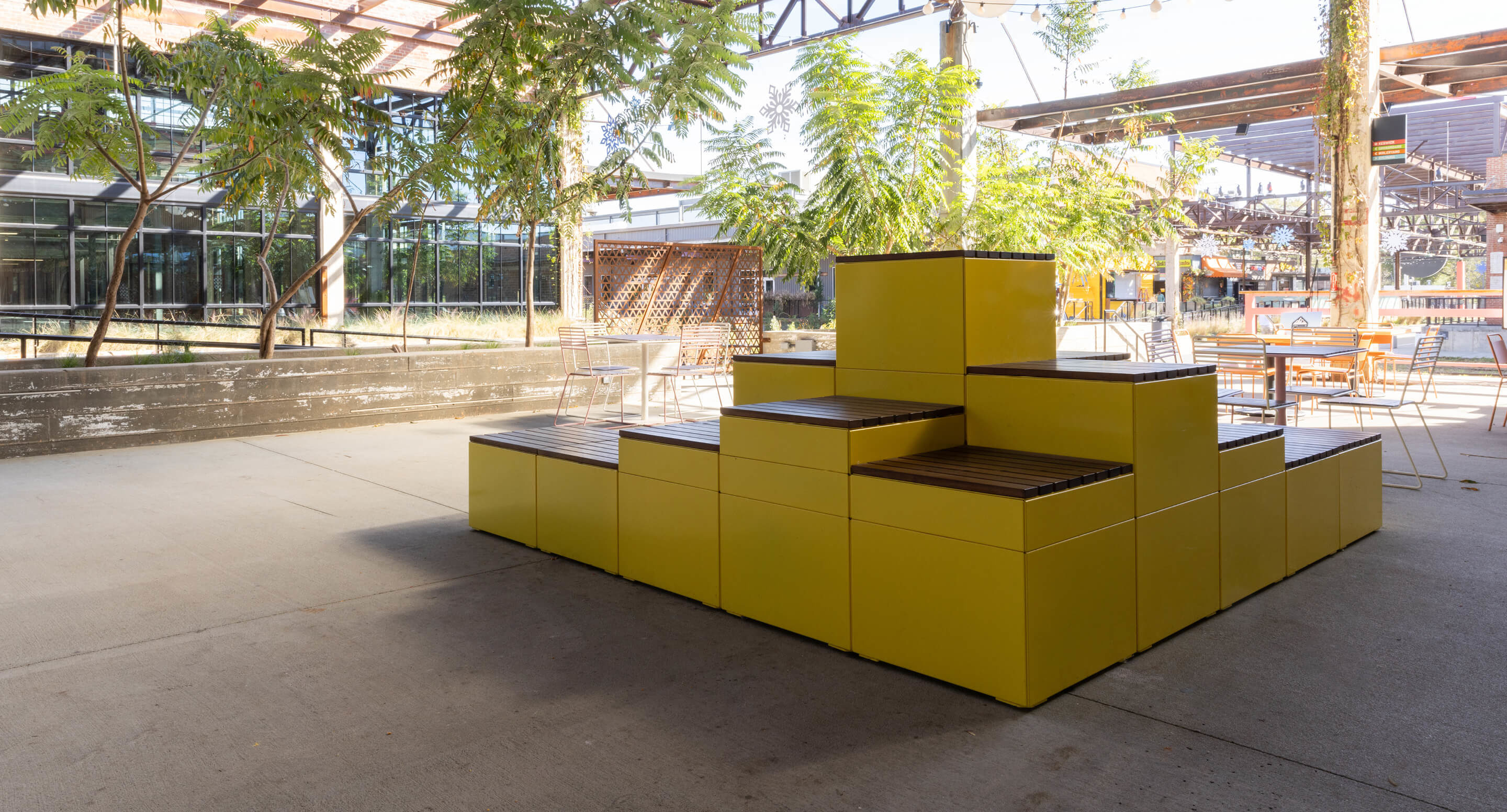maglin_pixel_5 A modular yellow bench structure with wooden seating is positioned outdoors in a shaded area with trees and scattered chairs.
