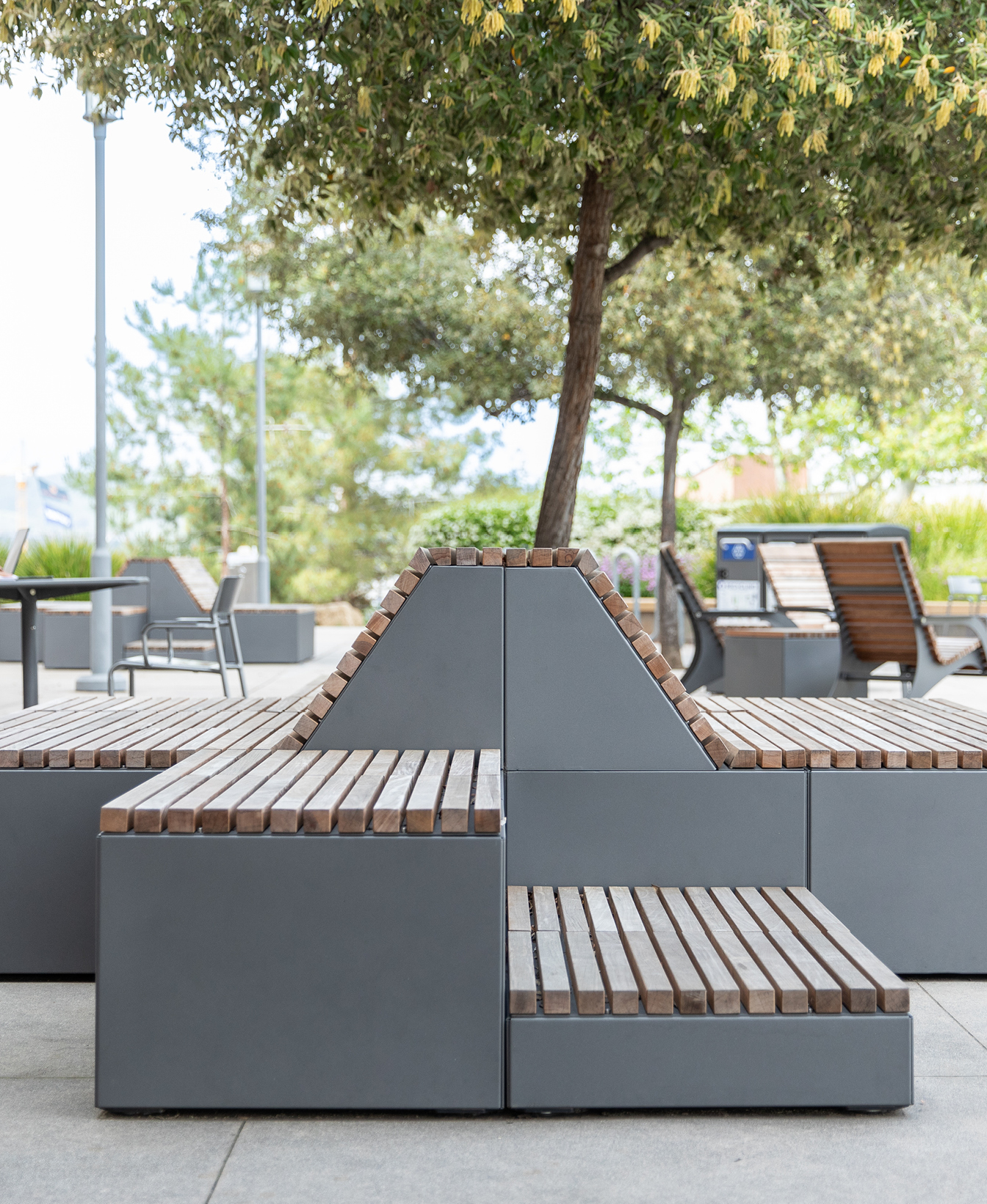 Pixel Seating: a modern outdoor bench with geometric design, crafted from gray metal and wooden slats, set beneath a leafy tree in a landscaped public space.