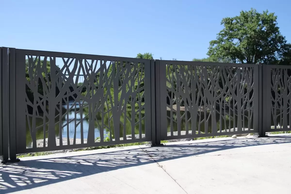 A metal fence with a tree branch pattern stands on a concrete surface, with water and trees visible in the background under a clear blue sky.