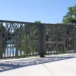 A metal fence with a tree branch pattern stands on a concrete surface, with water and trees visible in the background under a clear blue sky.