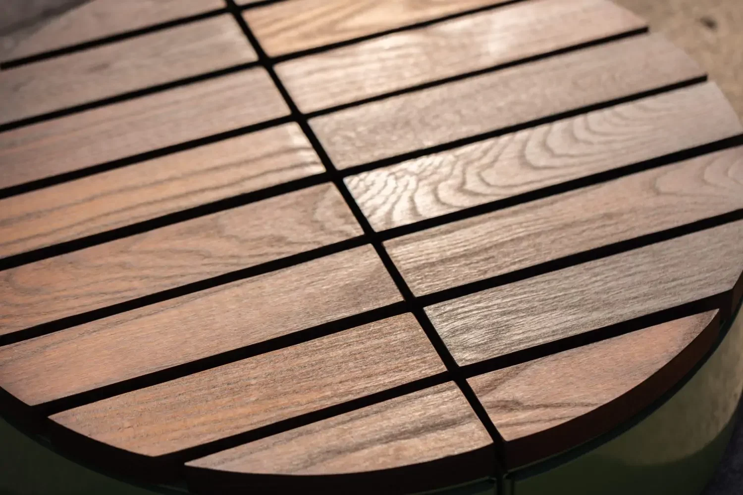 Close-up of a round wooden table with evenly spaced slats, showing the wood grain and sunlight reflecting off the surface.