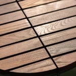 Close-up of a round wooden table with evenly spaced slats, showing the wood grain and sunlight reflecting off the surface.