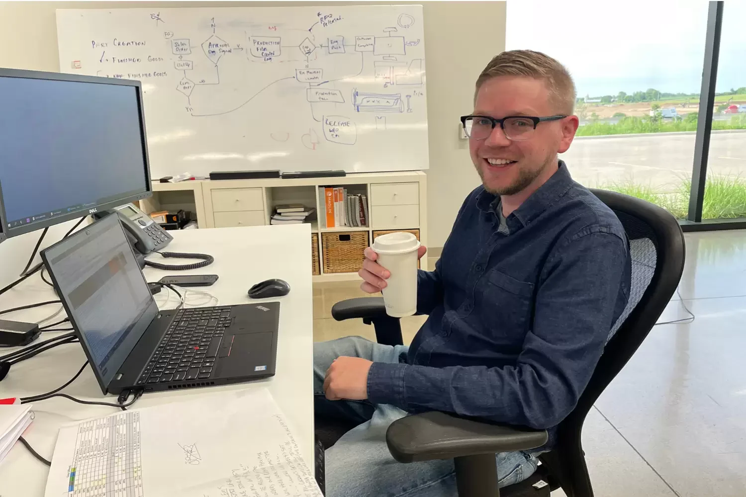 Oskar-Ahopelto A man sits at a Maglin desk with a laptop and paperwork, holding a coffee cup and smiling. A whiteboard with diagrams and notes is visible in the background.