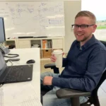 A man sits at a Maglin desk with a laptop and paperwork, holding a coffee cup and smiling. A whiteboard with diagrams and notes is visible in the background.