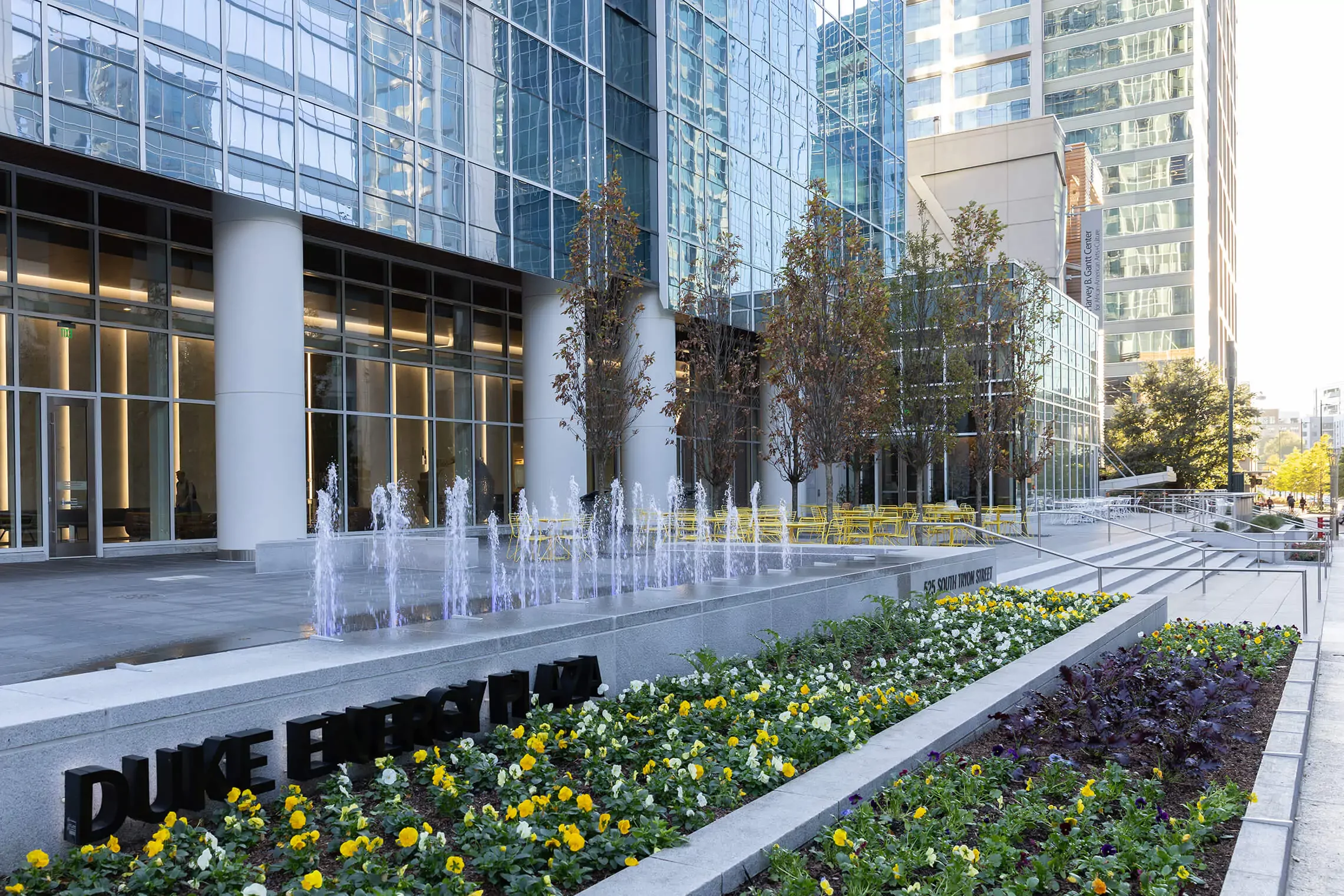 Entrance of Duke Energy Plaza with glass facade, flower beds, trees, water fountains, and nearby office buildings in an urban setting.
