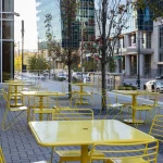 Empty yellow tables and chairs are arranged outdoors on a paved area near trees, with tall modern buildings and parked cars in the background.