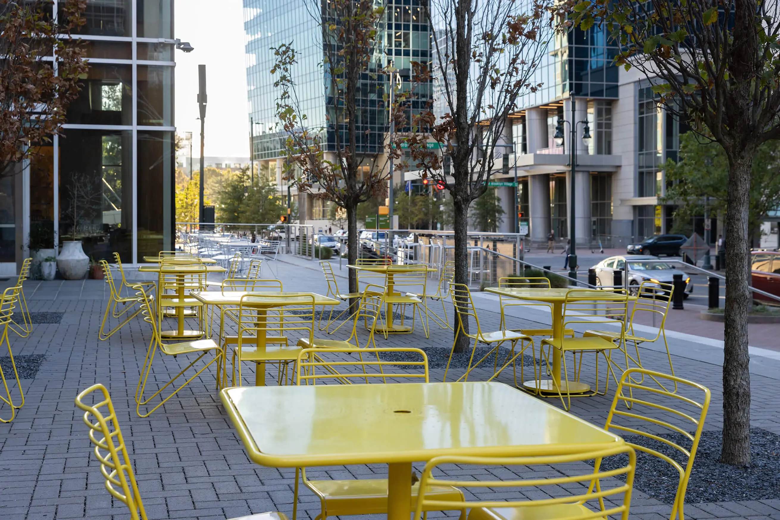 duke-energy-plaza-kontur-chairs-foro-square-tables-yellow-1 Espace extérieur avec plusieurs tables et chaises jaunes vides, entouré d'arbres, à côté d'une rue de la ville avec des bâtiments modernes en verre à la lumière du jour.