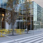 Outdoor seating area with yellow tables and chairs beside a modern glass building; a person walks down the steps in the foreground.