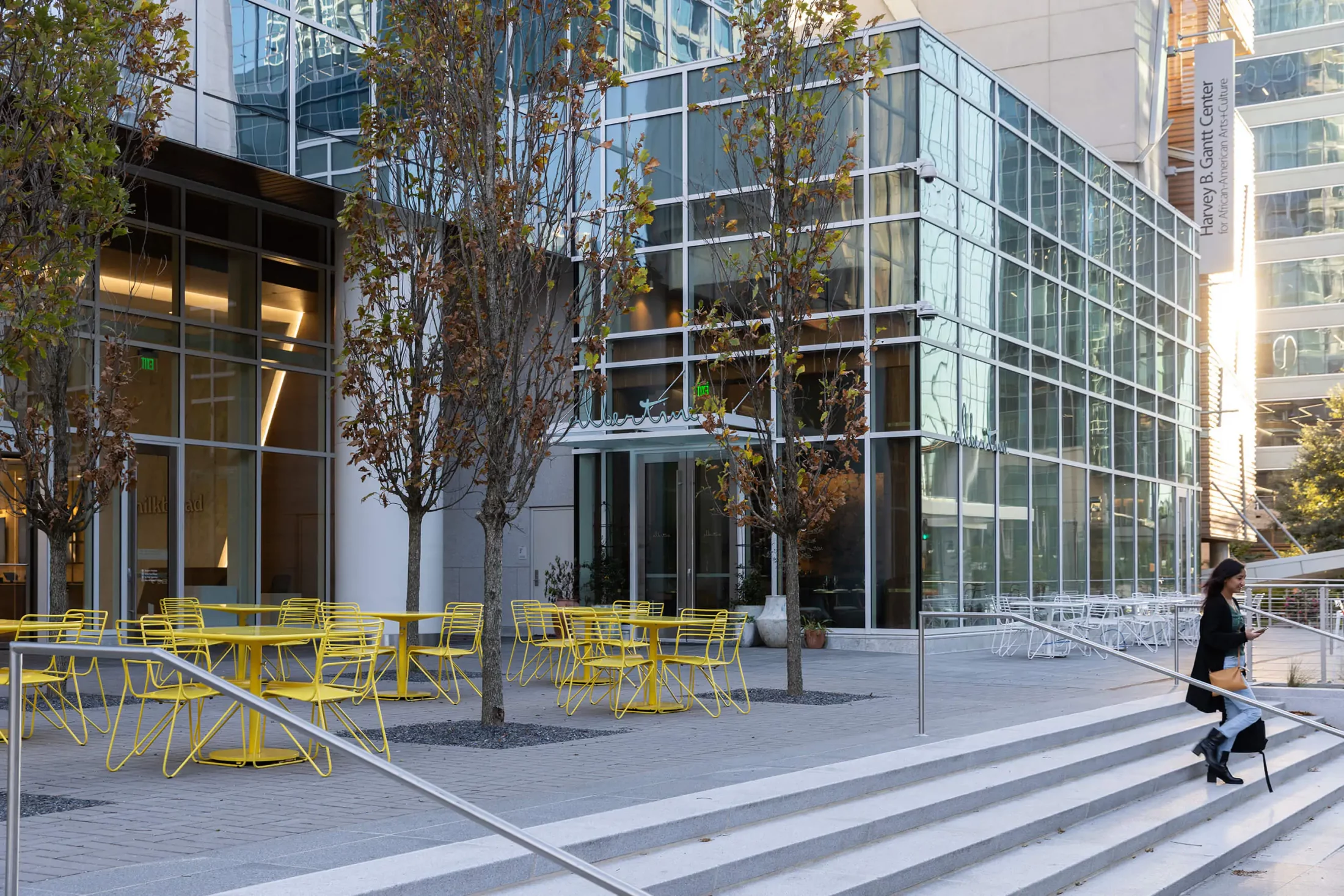 Outdoor seating area with yellow tables and chairs beside a modern glass building; a person walks down the steps in the foreground.