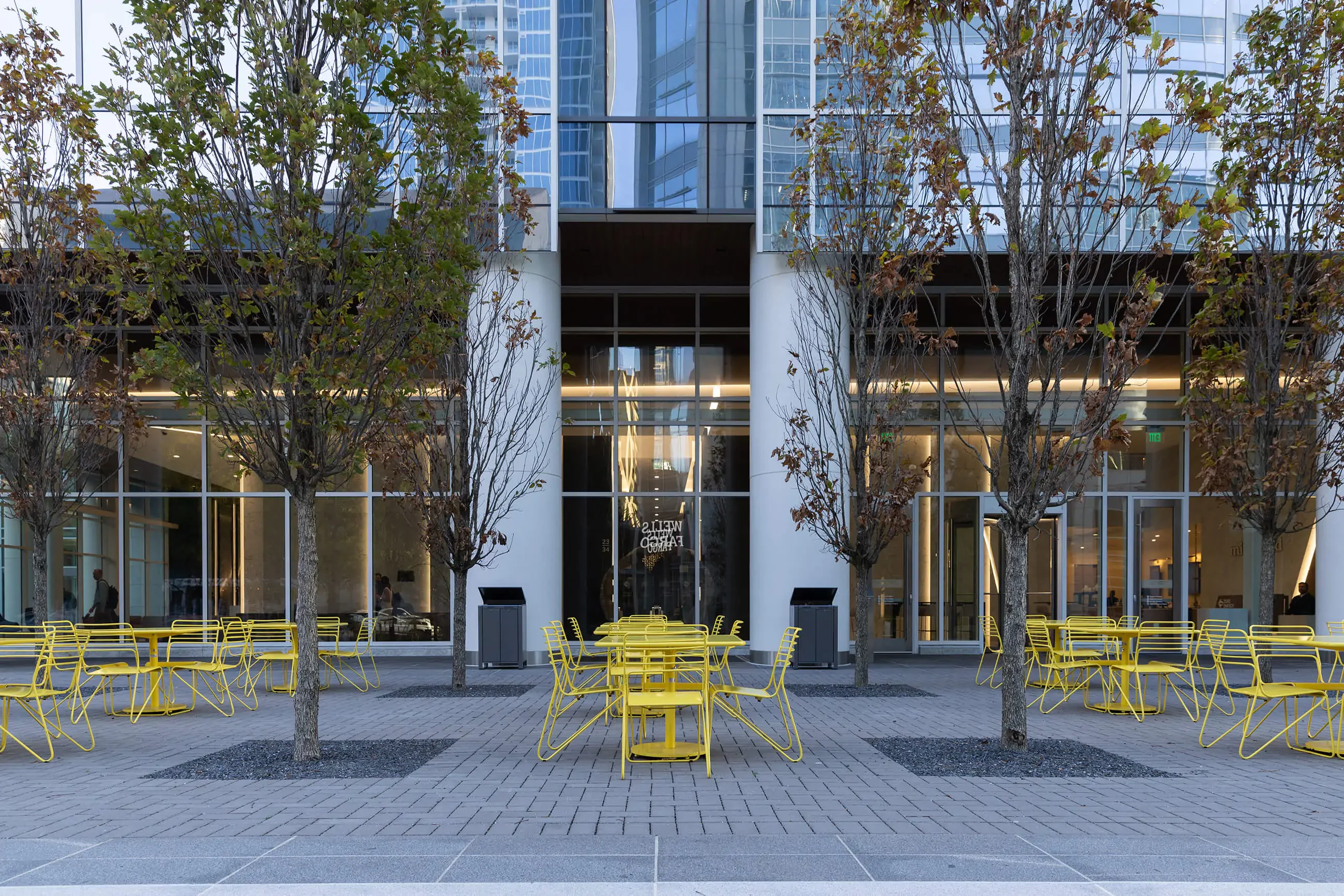 Outdoor café seating area with yellow tables and chairs, surrounded by trees, in front of a modern glass building.