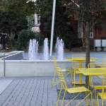 Des tables et des chaises en métal jaune sont disposées près d'un arbre et d'une petite fontaine sur une place en plein air, avec des gens qui marchent à l'arrière-plan.