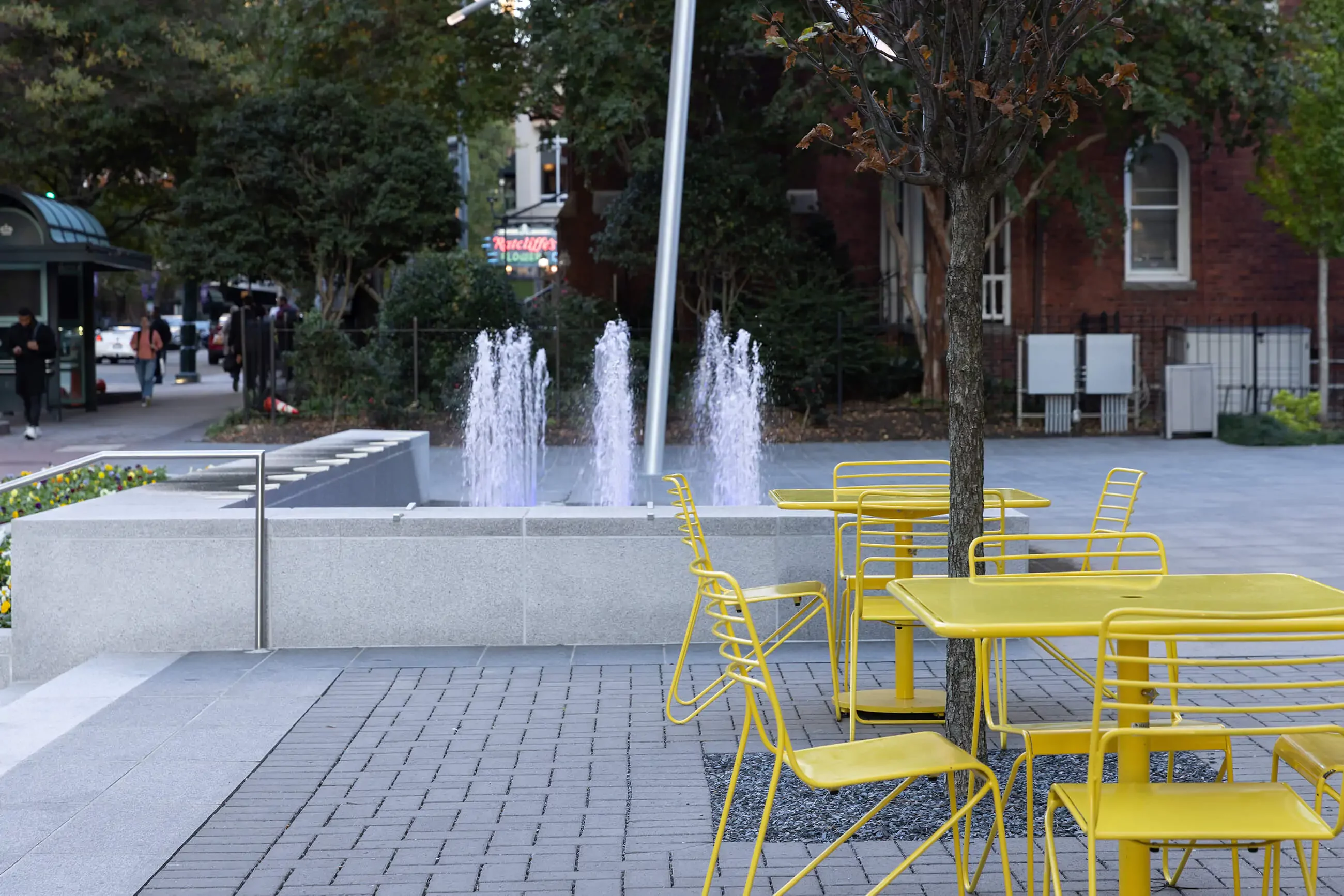 Outdoor plaza with yellow metal tables and chairs near a water fountain, surrounded by trees and buildings in the background.