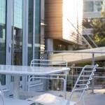 Table et chaises en métal blanc sur un patio extérieur, avec des bâtiments modernes en verre et de la verdure visibles à l'arrière-plan par une journée ensoleillée.
