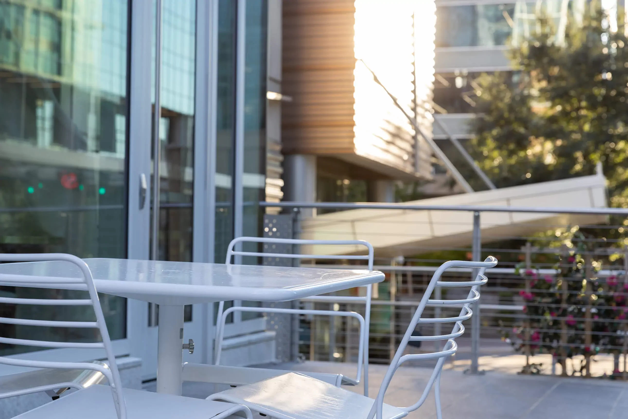 White metal table and chairs on an outdoor patio with modern buildings and glass windows in the background, sunlight reflecting off surfaces.