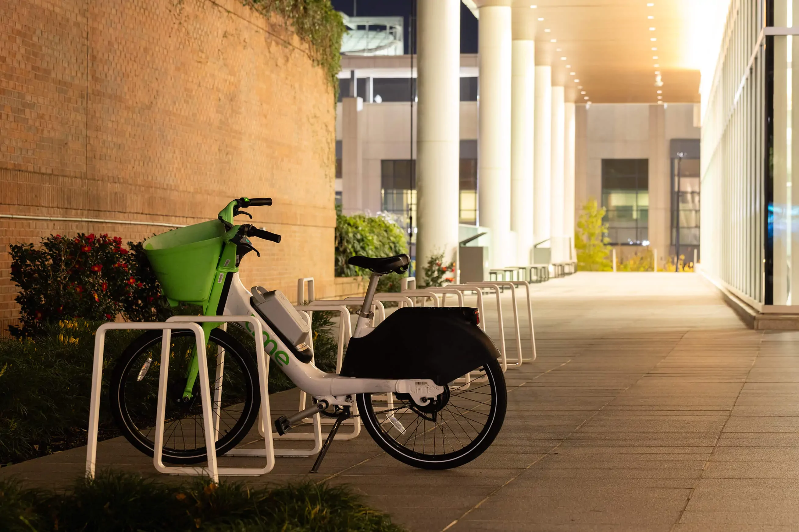 Two docked rental bikes are parked at a bike rack outside a modern building at night, illuminated by overhead lights.