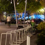 A mostly empty bike rack at night with one parked rental bike, a few people walking nearby, and cars driving past on a well-lit street.