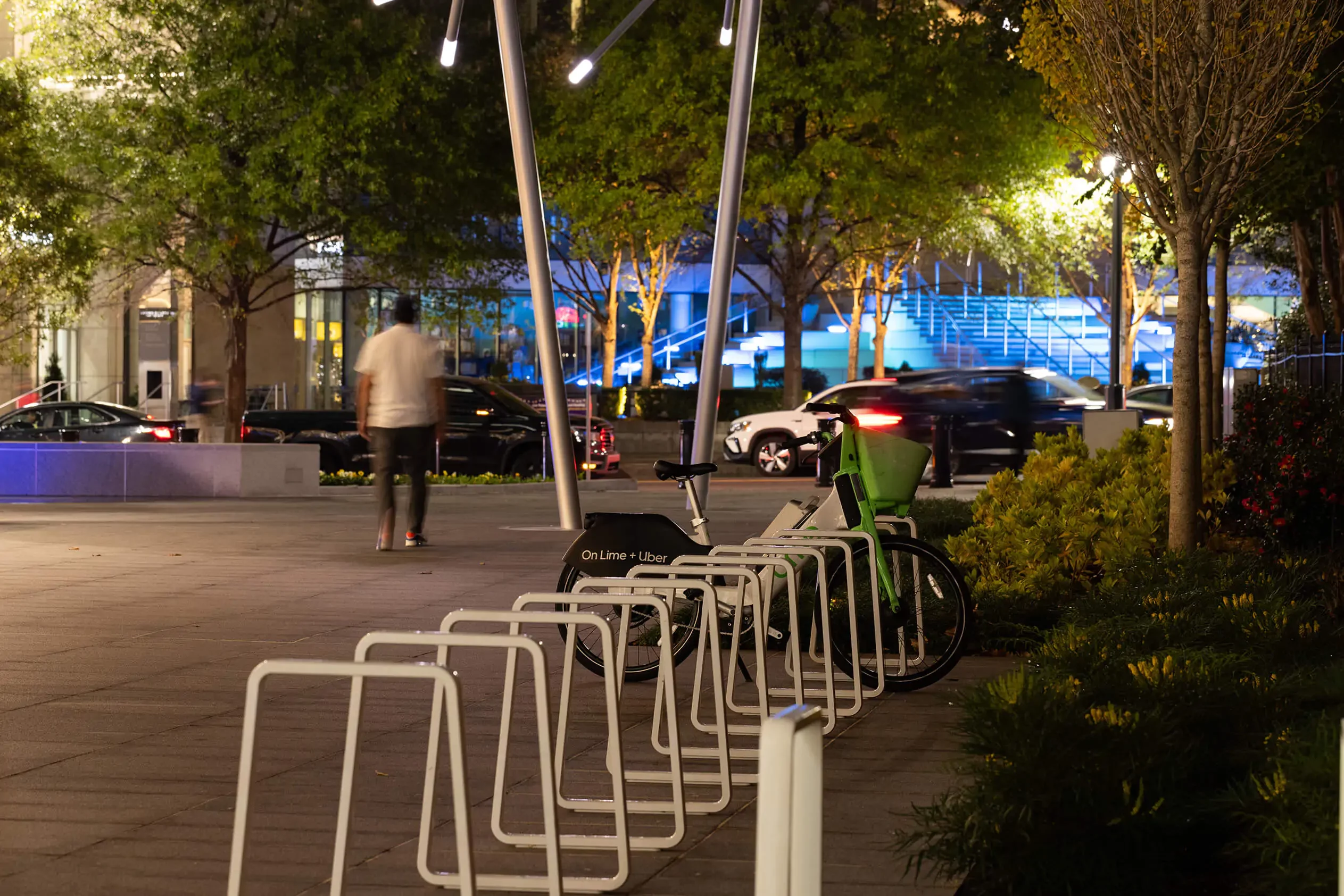 A mostly empty bike rack at night with one parked rental bike, a few people walking nearby, and cars driving past on a well-lit street.