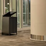 Une poubelle moderne noire à double compartiment pour les déchets et le recyclage, avec un couvercle ouvert, placée près d'une grande colonne et de fenêtres en verre sur un sol carrelé.