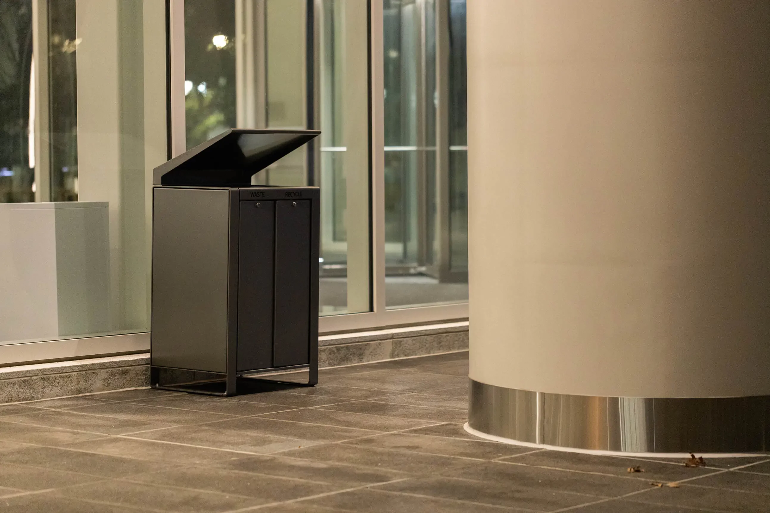 A black dual-compartment trash and recycling bin with an open lid is placed on a tiled floor next to a large cylindrical column in a modern indoor space.