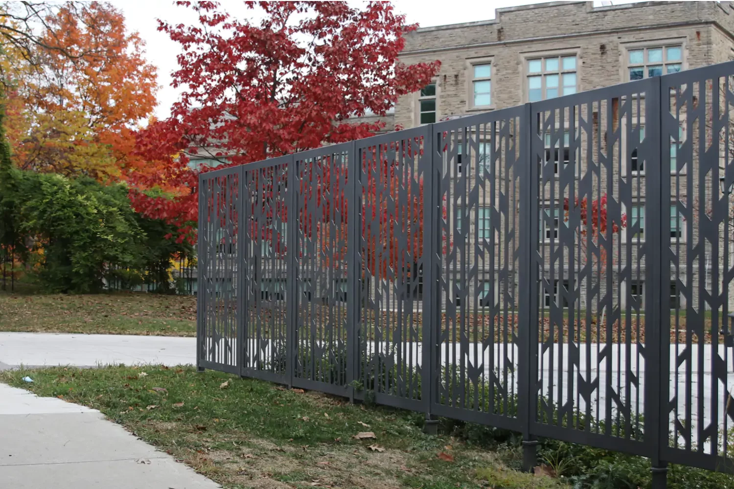 Decorative metal fence with an abstract pattern stands beside a sidewalk, with autumn trees and a multi-story brick building in the background.