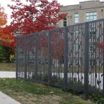 Decorative metal fence with an abstract pattern stands beside a sidewalk, with autumn trees and a multi-story brick building in the background.