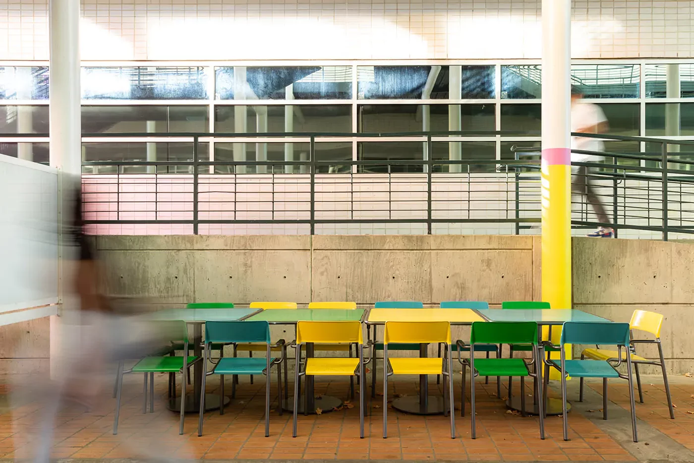 foro-chairs-tables-ncc-charlotte-colvard-breezeway-1 Plusieurs chaises vertes et jaunes sont disposées autour de tables dans un espace intérieur carrelé, avec des silhouettes floues qui passent, des grilles et des fenêtres à l'arrière-plan.
