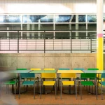 Plusieurs chaises vertes et jaunes sont disposées autour de tables dans un espace intérieur carrelé, avec des silhouettes floues qui passent, des grilles et des fenêtres à l'arrière-plan.