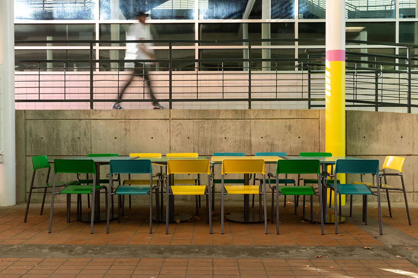 Rows of empty green and yellow chairs arranged around tables outdoors, with a person walking past in the background near a concrete wall and glass windows.