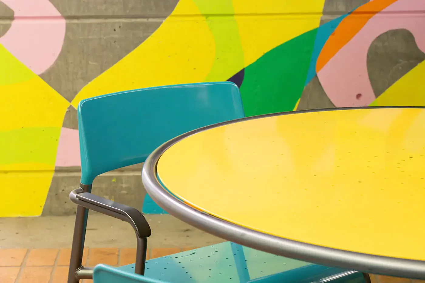 foro-chairs-tables-turquoise-yellow-ncc-charlotte-colvard-breezeway-1 Une table ronde jaune et des chaises sarcelles devant un mur en béton orné d'une peinture murale abstraite et colorée.