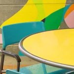 Une table ronde jaune et des chaises sarcelles devant un mur en béton orné d'une peinture murale abstraite et colorée.