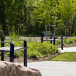 Three empty black benches line a curved concrete pathway surrounded by greenery and trees in a park setting near the renowned Canadian Canoe Museum.