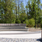 A curved wooden bench sits in a circular paved area surrounded by trees near the Canadian Canoe Museum, with a person riding a bicycle on a path in the background.