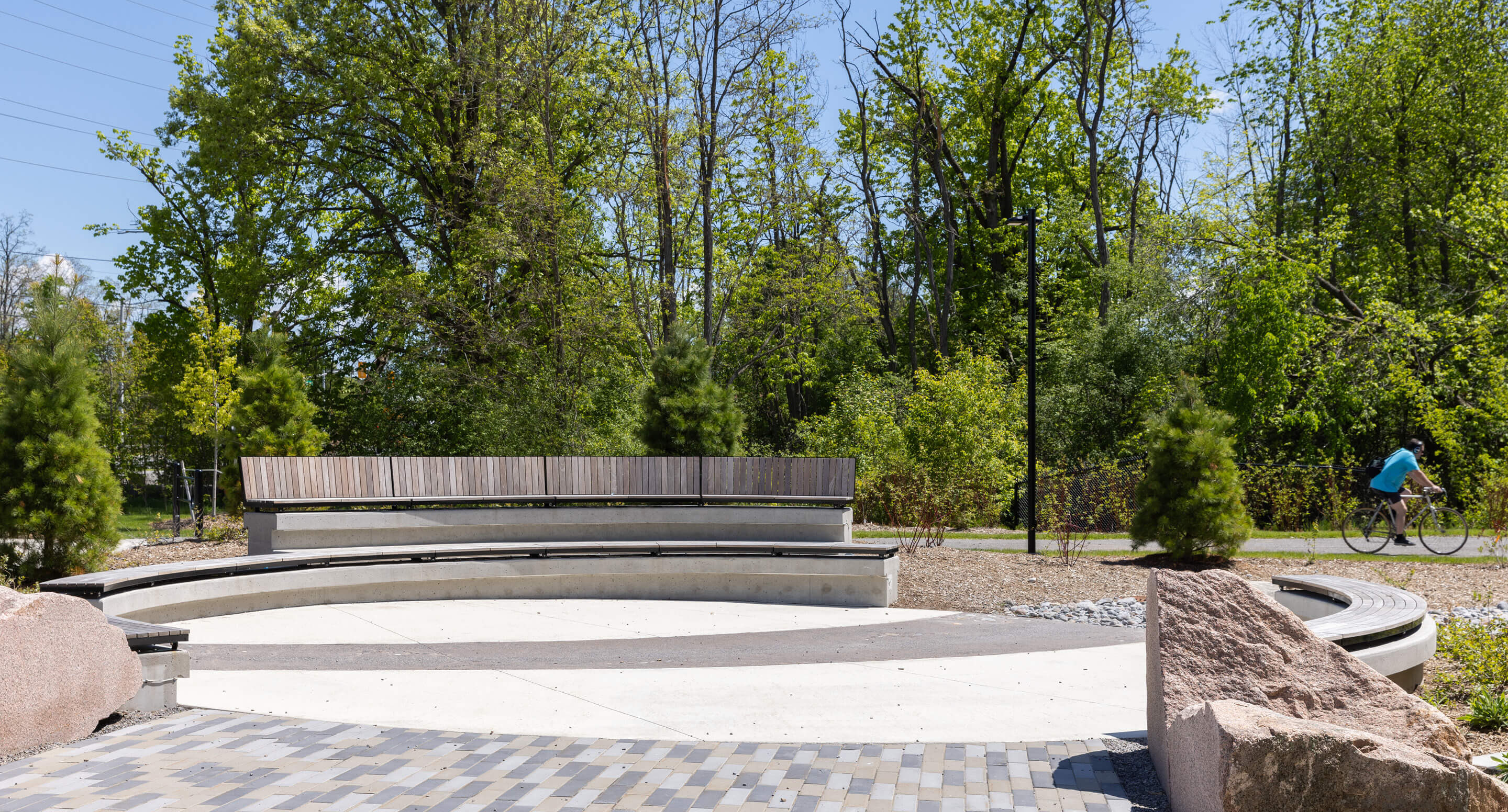 A curved wooden bench sits in a circular paved area surrounded by trees near the Canadian Canoe Museum, with a person riding a bicycle on a path in the background.