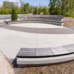 A circular outdoor seating area with curved concrete benches, surrounded by trees, shrubs, and a road in the background—perfect for visitors to relax near the Canadian Canoe Museum.