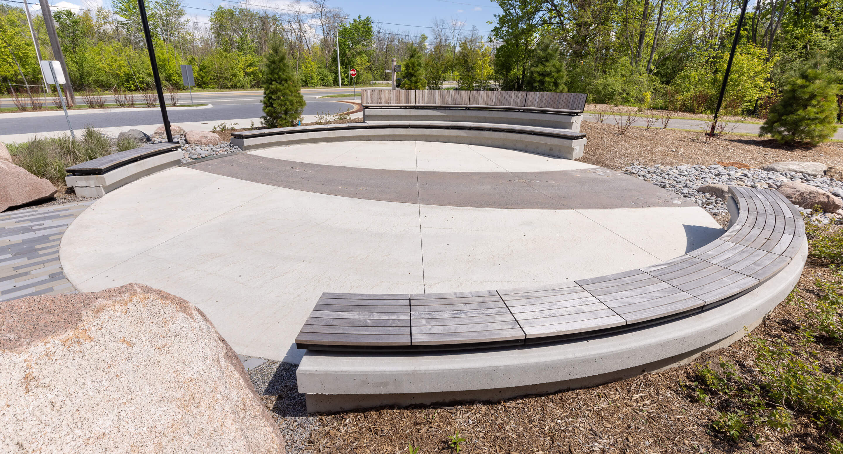 A circular outdoor seating area with curved concrete benches, surrounded by trees, shrubs, and a road in the background—perfect for visitors to relax near the Canadian Canoe Museum.