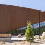 Modern building with angled rust-colored metal facade, large glass windows, and surrounding landscaping with rocks and young trees—home to the inspiring Canadian Canoe Museum.