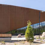 Modern building with angled rust-colored metal facade, large glass windows, and surrounding landscaping with rocks and young trees—home to the inspiring Canadian Canoe Museum.