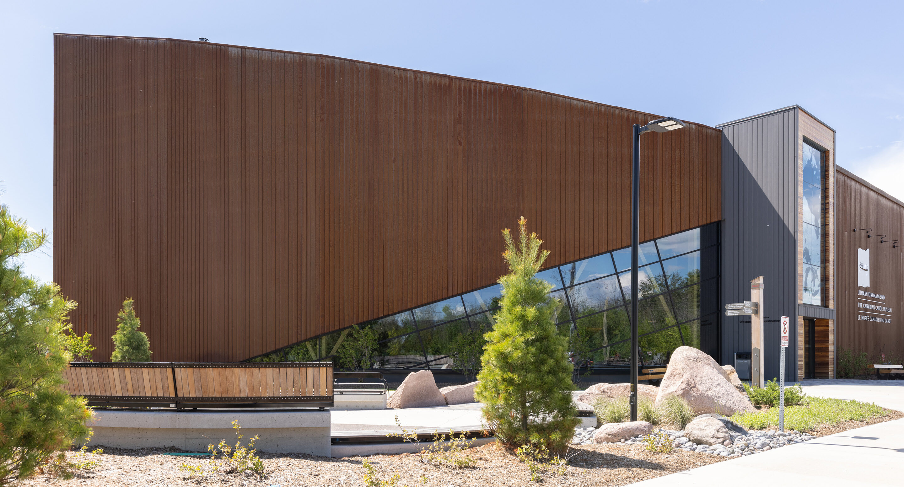 Modern building with angled rust-colored metal facade, large glass windows, and surrounding landscaping with rocks and young trees—home to the inspiring Canadian Canoe Museum.