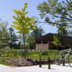 A bicycle is parked at a bike rack on a sunny day near the Canadian Canoe Museum, next to a wooden building surrounded by trees and green grass.