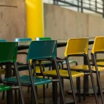 A row of green and yellow plastic chairs arranged around tables in an outdoor setting with concrete walls and metal railings.