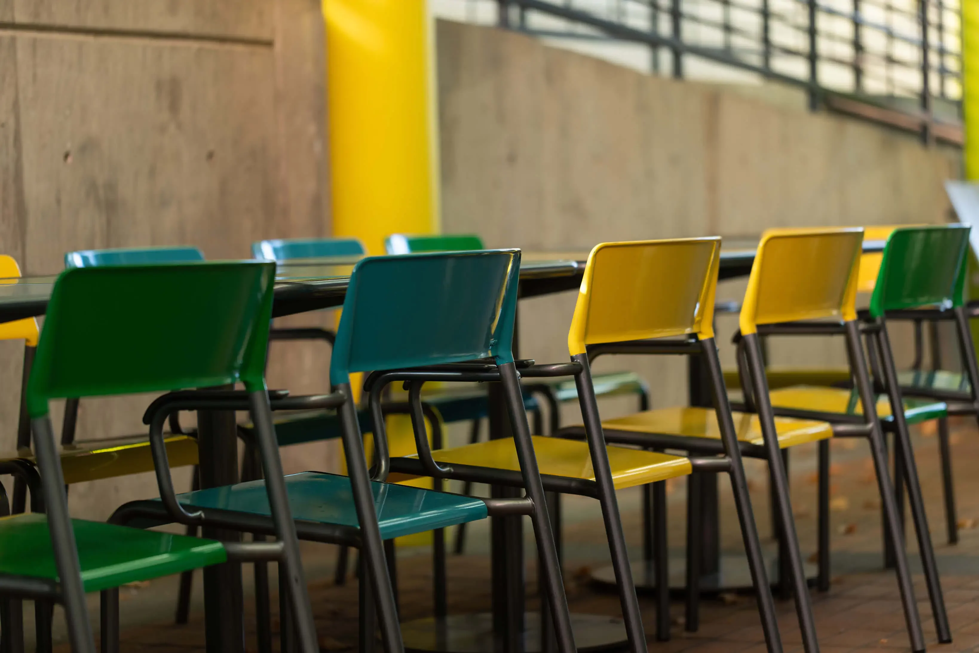 maglin-foro-chairs_1 Des rangées de chaises vides vertes et jaunes sont disposées à des tables dans un cadre extérieur avec un mur en béton en arrière-plan.